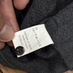 Puede incluir: Primer plano de una tela gris oscuro con una etiqueta adjunta. La etiqueta tiene el texto "NO. IQ-T90-113 Fabriqu&eacute; au Japon Made in Japan Y's bis Co Ltd" y un bot&oacute;n oscuro.