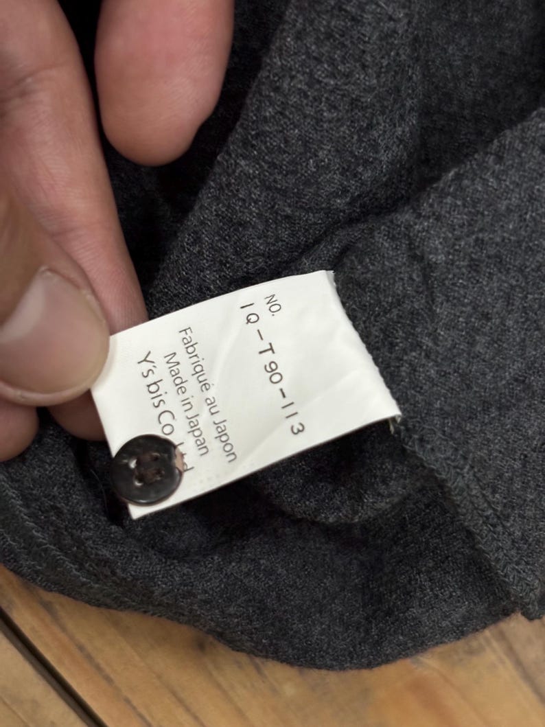 Puede incluir: Primer plano de una tela gris oscuro con una etiqueta adjunta. La etiqueta tiene el texto "NO. IQ-T90-113 Fabriqu&eacute; au Japon Made in Japan Y's bis Co Ltd" y un bot&oacute;n oscuro.