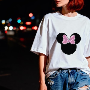May include: White t-shirt featuring a black Minnie Mouse head with a pink polka-dot bow. The shirt is a casual style, suitable for everyday wear. The design is centered on the front of the shirt.