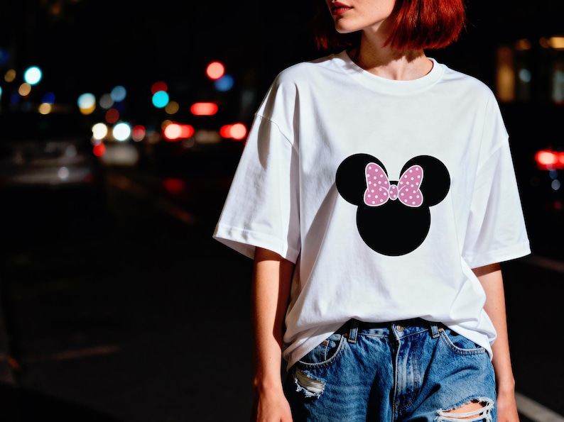 May include: White t-shirt featuring a black Minnie Mouse head with a pink polka-dot bow. The shirt is a casual style, suitable for everyday wear. The design is centered on the front of the shirt.