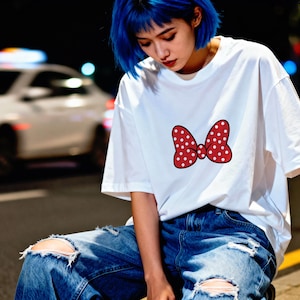 May include: White t-shirt featuring a red bow with white polka dots. The bow is centered on the front of the shirt. The person is wearing ripped blue jeans. The background is a city street at night.