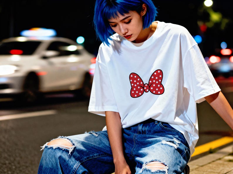 May include: White t-shirt featuring a red bow with white polka dots. The bow is centered on the front of the shirt. The person is wearing ripped blue jeans. The background is a city street at night.