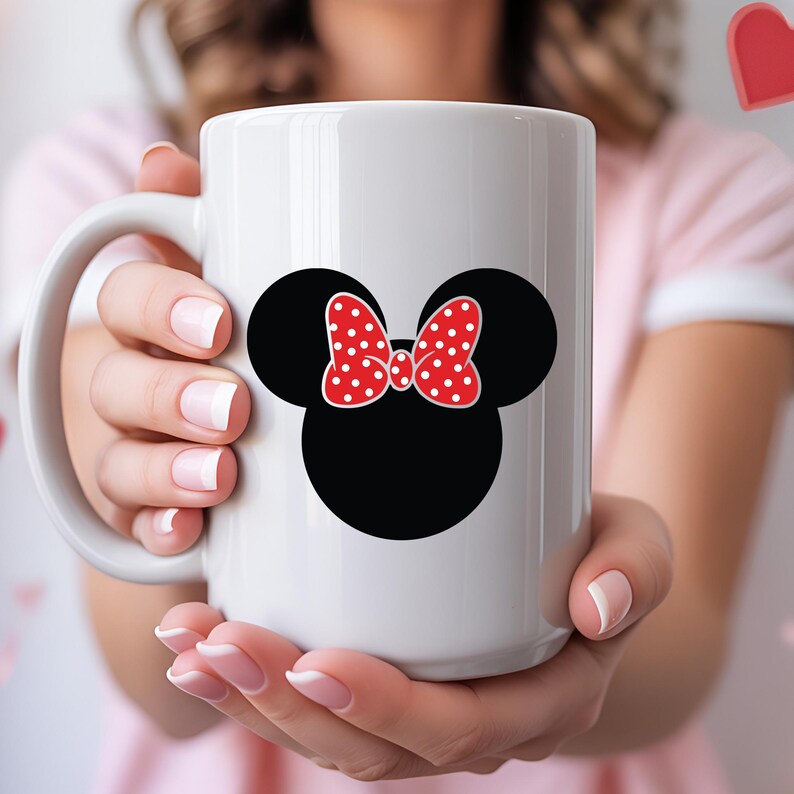 May include: White ceramic mug featuring a black silhouette of mouse ears with a red bow with white polka dots. The mug is held by a person with pink nail polish.