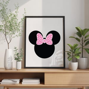 May include: Black framed wall art featuring a silhouette of a mouse head with a pink polka dot bow. The artwork is displayed on a wooden console table with decorative plants in white and gray pots. The background is a neutral wall.