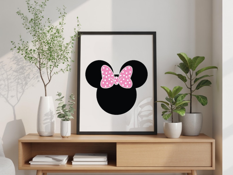 May include: Black framed wall art featuring a silhouette of a mouse head with a pink polka dot bow. The artwork is displayed on a wooden console table with decorative plants in white and gray pots. The background is a neutral wall.