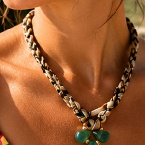 Può includere: Una collana con un cordino in tessuto intrecciato in un motivo animalier nero, bianco e marrone chiaro. Il ciondolo è composto da tre pietre verdi lisce a forma di goccia disposte a grappolo, impreziosite da piccole perline in metallo dorato.