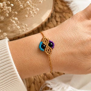 Può includere: Un braccialetto macramè fatto a mano con un design quadrato centrale con accenti neri, viola e turchese. Il braccialetto è realizzato con un cordoncino color oro ed è esposto su un polso.