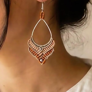 Peut inclure: Une boucle d'oreille en forme de goutte d'eau avec un tressage macramé complexe dans des tons orange brûlé et crème. Le design comprend une armature en métal argenté, une petite perle argentée au centre et un crochet argenté pour l'oreille.