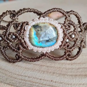 May include: A macrame bracelet featuring a central oval cabochon stone with iridescent blue and green flashes. The stone is framed by a light pink knotted border, surrounded by intricate brown cord work with small embedded silver-toned metallic beads.