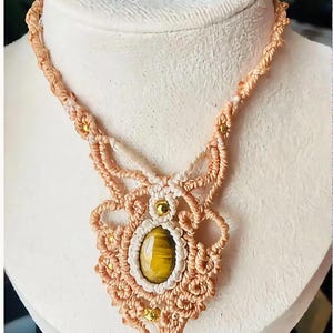 May include: A macrame necklace featuring a central oval tiger's eye gemstone in a golden-brown hue. The pendant is crafted from light peach-colored cord with intricate knotted patterns, accented by small metallic gold beads throughout the design.