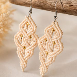 Pode incluir: Um par de brincos de macramé na cor creme com um padrão geométrico de losangos atados. Cada brinco é acentuado com pequenas contas redondas em tom dourado integradas na estrutura do cordão e pendurado num gancho de metal prateado.