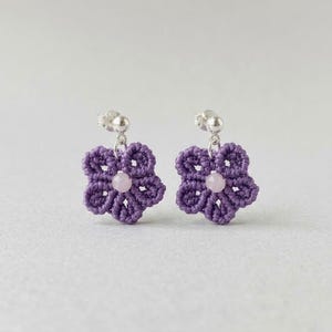 Könnte beinhalten: Ein Paar Ohrstecker mit violetten Makramee-Blumenmotiven. Jede Blume ist aus geknotetem Garn gefertigt und mit einer kleinen, runden, blassrosa Perle in der Mitte versehen. Die Ohrringe sind an silberfarbenen Metallsteckern befestigt.