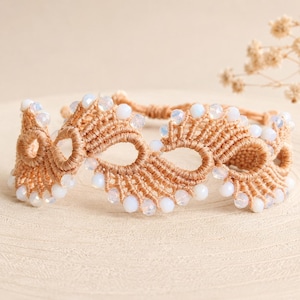 May include: A macrame bracelet crafted from tan cord featuring an intricate wave-like pattern. The design is accented with small, translucent white beads embedded along the edges of the woven structure, creating a delicate and textured appearance.