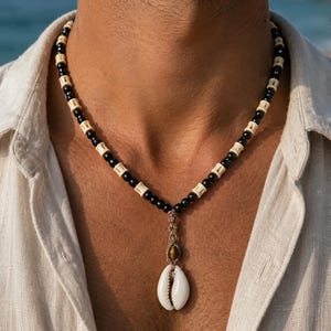 Puede incluir: Un collar de cuentas con una alternancia de cuentas esféricas negras y espaciadores cilíndricos de color crema. El colgante central consta de una cuenta de ojo de tigre marrón unida a una concha de cauri blanca, suspendida por un cordón trenzado beige.