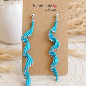May include: A pair of long, spiral-shaped earrings made of bright turquoise knotted thread, accented with small silver-coloured beads throughout. The earrings are attached to a rectangular brown card with the text Handmade with love printed on it.