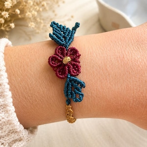 Può includere: Un braccialetto macramè fatto a mano con un fiore bordeaux con centro dorato e foglie turchesi. Il braccialetto è collegato con macramè color oro, un gioiello unico.