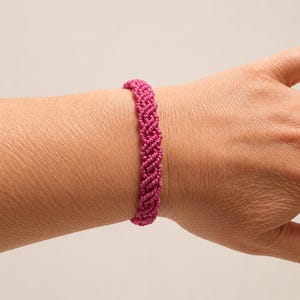 Può includere: Un braccialetto color magenta vivace realizzato con cordino annodato in modo intricato. Il design presenta un motivo intrecciato ripetitivo creato tramite tecniche di macramè, formando una fascia continua e testurizzata attorno al polso.