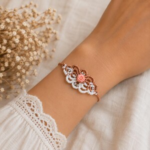 Può includere: Un bracciale in macramè con un ciondolo centrale a forma di rosa color pesca, intricati motivi a cordoncino intrecciato marrone e bianco e piccole perline metalliche argentate integrate nel design, fissato con un sottile cordino marrone.