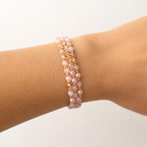 Può includere: Un bracciale a più fili caratterizzato da tre file di piccole perline sfaccettate rosa chiaro, intrecciate insieme con un cordoncino sottile di colore chiaro. Le perline hanno un riflesso tenue e sono disposte in un motivo uniforme.