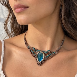 Può includere: Una collana in macramè con una pietra di labradorite centrale a forma di goccia con riflessi blu iridescenti. La pietra è incorniciata da un intreccio di cordino marrone e decorata con piccole perline color ottanio e minuscole sfere argentate.