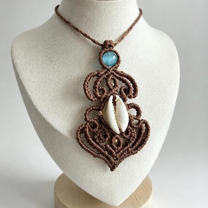 Può includere: Una collana in macramè marrone con una conchiglia di ciprea bianca centrale, una pietra preziosa rotonda azzurra sulla parte superiore e piccole perline dorate integrate nell'intricato design a nodi, su un cordino intrecciato marrone.