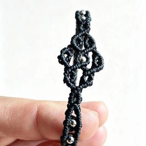 May include: A black macrame bracelet featuring intricate knotted patterns and small, polished silver-colored metallic beads integrated throughout the design. The bracelet displays a central decorative element with a symmetrical, open-work knotted structure.