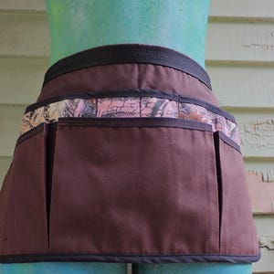 May include: A brown waist apron featuring a horizontal band of pink and brown camouflage pattern across the top section. The apron includes three large, deep pockets across the front and is finished with black trim along the edges and waistband.