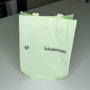Puede incluir: Una bolsa tote verde claro con el nombre de la marca "lululemon" impreso en negro. La bolsa tiene un cierre de botón a presión y dos asas pequeñas. El logotipo de lululemon está en el lado izquierdo.