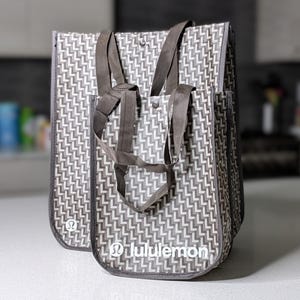May include: Two gray and white patterned tote bags with brown handles. The bags have a repeating L-shaped design. The brand name "lululemon" is printed in white on the front of the smaller bag.