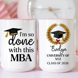 Puede incluir: Dos tazas de cerámica blancas con asas y bordes rosas. Una taza dice "I'm so done with this MBA" y presenta un birrete. La otra taza tiene un birrete, un pergamino y el nombre "Evelyn".