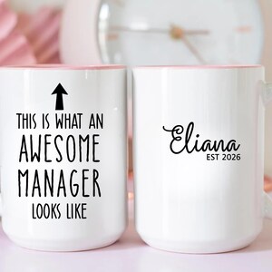 Puede incluir: Taza de cerámica blanca con asa e interior rosa. Un lado dice "THIS IS WHAT AN AWESOME MANAGER LOOKS LIKE" con una flecha. El otro lado tiene el nombre "Eliana" y "EST 2026".
