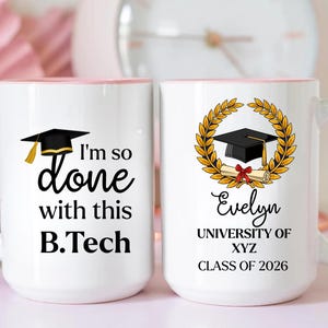 May include: Two white ceramic mugs with pink handles and rims. One mug reads "I'm so done with this B.Tech" with a graduation cap graphic. The other mug features a graduation cap, laurel wreath, and the name "Evelyn", university details, and "Class of 2026".