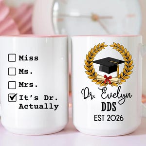 May include: White ceramic mug with a pink handle and rim. One side has text options for titles, with "It's Dr. Actually" checked. The other side features a graduation cap, diploma, and the text "Dr. Evelyn DDS EST 2026".
