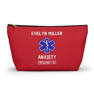 Puede incluir: Una bolsa roja con cremallera con el texto "EVELYN MILLER" encima de un símbolo azul de la Estrella de la Vida y las palabras "ANXIETY EMERGENCY KIT". La bolsa tiene una cremallera negra y un tirador dorado.