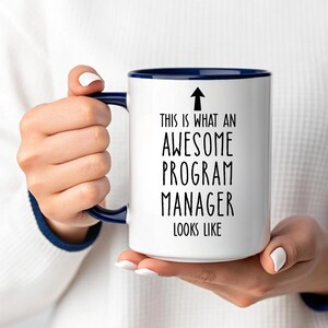May include: White ceramic mug with a navy blue interior and handle. The mug features the text "THIS IS WHAT AN AWESOME PROGRAM MANAGER LOOKS LIKE" in black, with a black arrow pointing upwards.