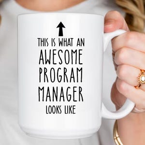 May include: White ceramic mug with the text "THIS IS WHAT AN AWESOME PROGRAM MANAGER LOOKS LIKE" in black. An arrow points upwards above the text. The mug has a large handle.