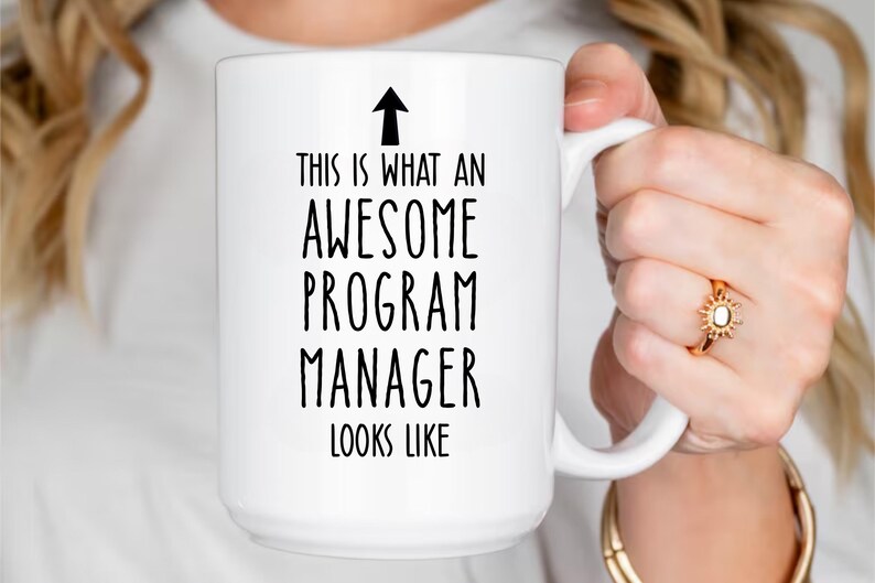May include: White ceramic mug with the text "THIS IS WHAT AN AWESOME PROGRAM MANAGER LOOKS LIKE" in black. An arrow points upwards above the text. The mug has a large handle.