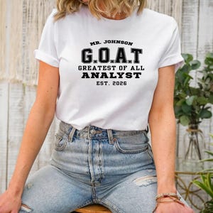 Può includere: Una t-shirt bianca a maniche corte con testo nero che recita MR. JOHNSON G.O.A.T. GREATEST OF ALL ANALYST EST. 2026. La maglietta ha un girocollo ed è abbinata a jeans in denim azzurro slavato con strappi visibili sul tessuto.