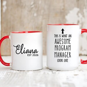 May include: Two white ceramic mugs with red handles and rims. One mug has the name "Eliana" in cursive with "EST 2026" below it. The other mug says "THIS IS WHAT AN AWESOME PROGRAM MANAGER LOOKS LIKE" with an arrow pointing up.