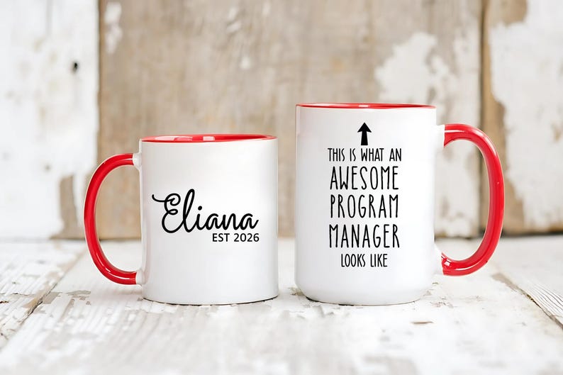 May include: Two white ceramic mugs with red handles and rims. One mug has the name "Eliana" in cursive with "EST 2026" below it. The other mug says "THIS IS WHAT AN AWESOME PROGRAM MANAGER LOOKS LIKE" with an arrow pointing up.
