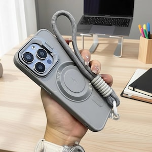 May include: Gray phone case with a magnetic charging circle and a gray lanyard. The case has a camera lens protector and a white loop for the lanyard. The phone case is held in a person's hand.