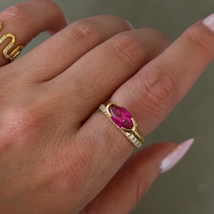 Può includere: Un anello d'oro con una grande gemma ovale color magenta. La fascia è inoltre ornata da piccole gemme trasparenti. Un altro anello d'oro con un design a serpente è visibile. Gioielli.
