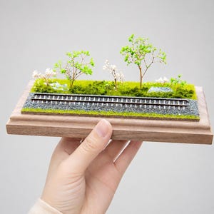 May include: Miniature railway diorama featuring a track with gravel, green grass, and small trees with green and white foliage. The diorama is set on a wooden base and held in a hand.