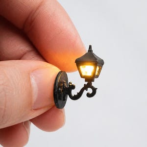 May include: A miniature black wall-mounted lantern with a decorative scroll arm and a glowing yellow light. The small lamp features a classic hexagonal housing with clear panels, designed for scale models or dollhouse lighting applications.