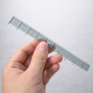 May include: A miniature grey fence model with vertical and horizontal lines, held by a hand against a white background. The fence has evenly spaced vertical supports and a mesh-like appearance.