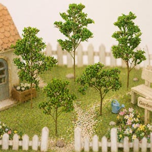 May include: Miniature garden scene with several small green trees, a white picket fence, and a small house. A bench and a sign that says "Garden" are also visible. The scene is set on a green grassy area with a stone pathway.