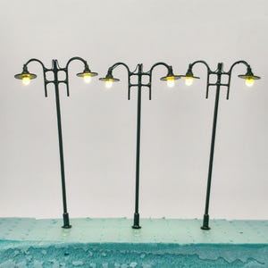 Può includere: Miniature lampioni verde scuro con lampadine gialle. Le lampade hanno un design classico con un braccio curvo e un supporto decorativo. I lampioni sono disposti in fila, perfetti per plastici ferroviari.
