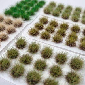 May include: A selection of miniature model grass tufts in shades of green and beige. The tufts are presented on clear plastic sheets, suitable for model making or diorama projects. The image is well-lit, highlighting the texture and colour variations of the grass.