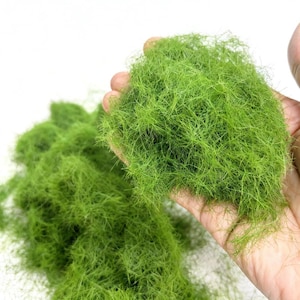May include: A handful of bright green model railroad foliage, resembling fine grass, held against a white background. The texture is dense and fibrous, with a vibrant, naturalistic appearance. Additional clumps of the same material are visible.