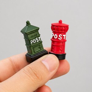 May include: Two miniature post boxes, one green and one red, are held in a hand. The green post box is rectangular, while the red one is cylindrical. Both have the word "POST" in white lettering.